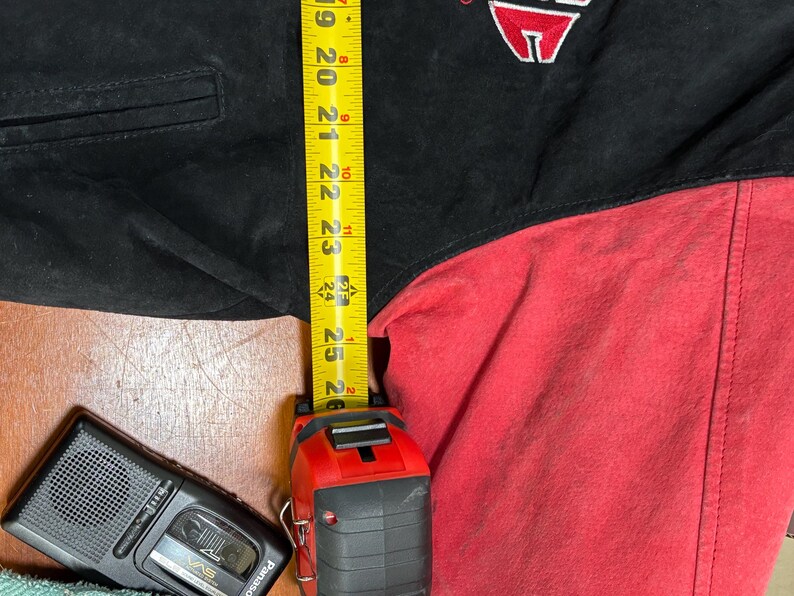 Puede incluir: Chaqueta negra y roja con un logotipo en la parte superior izquierda del pecho. Una cinta m&eacute;trica amarilla muestra una longitud de aproximadamente 63,5 cm. Un peque&ntilde;o reproductor de casetes negro est&aacute; en la esquina inferior izquierda.