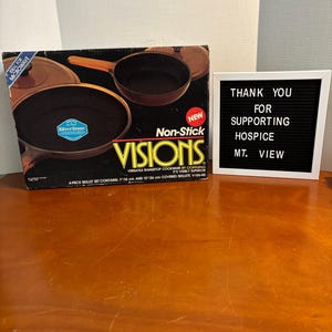 Könnte beinhalten: Ein neues Visions Antihaft-Kochgeschirr-Set mit zwei Bratpfannen, einer 18 cm und einer 26 cm, wird in seiner Originalverpackung präsentiert. Die Box zeigt Bilder des Kochgeschirrs und den Text "IDEAL FOR MICROWAVE". Ein Schild lautet "THANK YOU FOR SUPPORTING HOSPICE MT. VIEW."