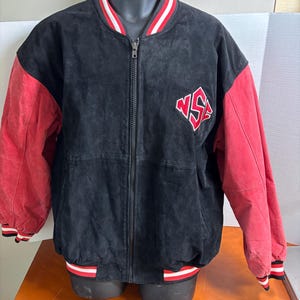 May include: Black suede jacket with red sleeves and a red, white, and black striped trim. The jacket features a full zipper and a red and white logo on the chest. The jacket is displayed on a black mannequin.