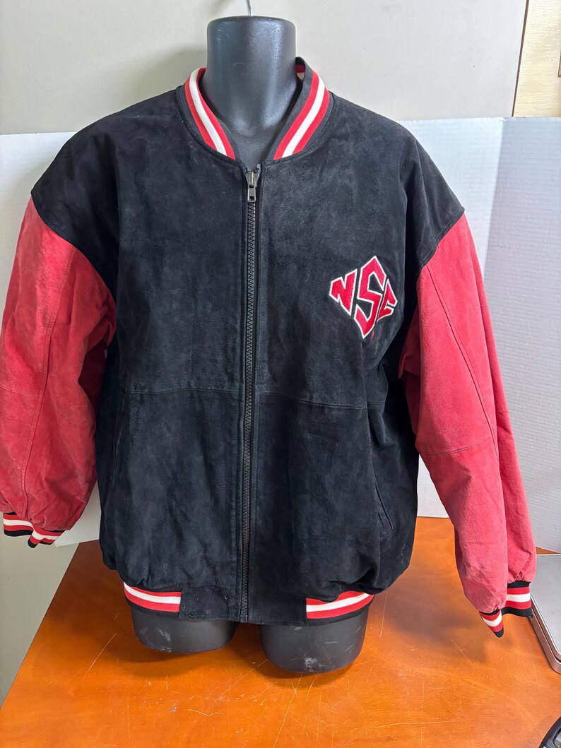 Puede incluir: Chaqueta de ante negra con mangas rojas y ribete a rayas rojas, blancas y negras. La chaqueta tiene una cremallera completa y un logotipo rojo y blanco en el pecho. La chaqueta se muestra en un maniqu&iacute; negro.
