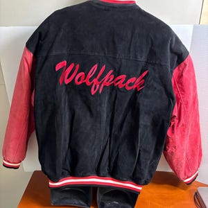 Puede incluir: Chaqueta universitaria negra y roja con la palabra "Wolfpack" bordada en rojo en la espalda. La chaqueta tiene mangas rojas y un cuerpo negro. El cuello y los pu&ntilde;os tienen rayas blancas y rojas.