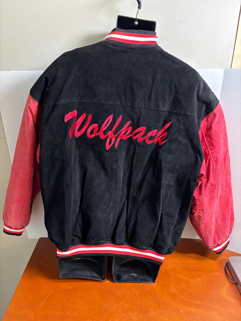 Puede incluir: Chaqueta universitaria negra y roja con la palabra "Wolfpack" bordada en rojo en la espalda. La chaqueta tiene mangas rojas y un cuerpo negro. El cuello y los pu&ntilde;os tienen rayas blancas y rojas.