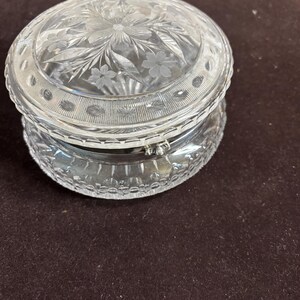 May include: A round, clear glass trinket box featuring an intricate floral etching on the lid and a decorative band of repeating oval patterns. The base and lid are joined by a silver-toned metal hinge, showcasing a polished, transparent finish.