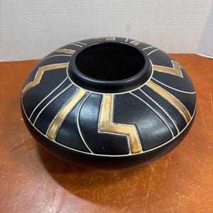 Könnte beinhalten: Eine niedrige, abgerundete schwarze Keramikvase mit einer breiten kreisförmigen Öffnung. Die Außenfläche ist mit einem sich wiederholenden geometrischen Muster aus goldenen Zickzacklinien und feinen weißen Umrandungen verziert.