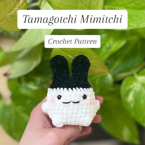 May include: A handmade crochet Mimitchi character from Tamagotchi, featuring a white body, black ears, and a smiling face with rosy cheeks. The text "Tamagotchi Mimitchi" and "Crochet Pattern" are displayed above the character.