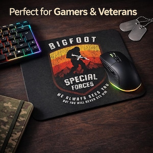 May include: Black gaming mouse and mousepad with a Bigfoot Special Forces design. The mousepad features a Bigfoot silhouette with the text "He Always Sees You But You Will Never See Him". A keyboard and dog tags are also visible.