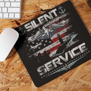 May include: Black mousepad with a distressed American flag design, featuring the text "SILENT SERVICE." The design includes anchors, stars, and aquatic illustrations. A white mouse and keyboard are visible.
