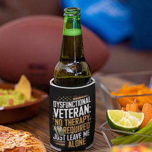 May include: A green glass beer bottle in a black koozie with the text "Dysfunctional Veteran: No Therapy Required, Just Leave Me Alone." The koozie has a patriotic design. The bottle is on a wooden table with food.