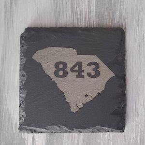 May include: Black slate coaster with a gray outline of the state of South Carolina and the number 843 printed in black.