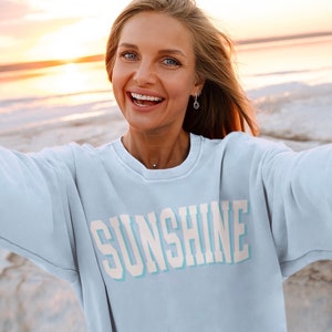 Peut inclure: Une femme portant un sweat-shirt bleu clair avec le mot "SUNSHINE" imprim&eacute; en blanc sur le devant. Elle sourit et regarde la cam&eacute;ra. L'arri&egrave;re-plan est une plage avec une &eacute;tendue d'eau et un coucher de soleil.