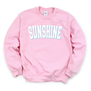 Peut inclure: Un sweat-shirt rose avec le mot "SUNSHINE" imprim&eacute; en blanc avec un contour bleu clair.