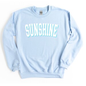 Peut inclure: Un sweat-shirt bleu clair avec le mot "SUNSHINE" imprim&eacute; en blanc avec un contour bleu clair.