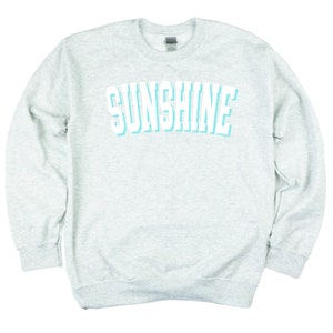 Peut inclure: Un sweat-shirt gris chin&eacute; avec le mot "SUNSHINE" imprim&eacute; en blanc avec un contour bleu clair.