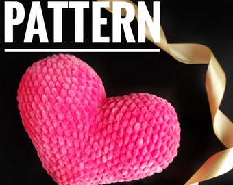 Puede incluir: Una almohada en forma de corazón rosa con una superficie texturizada. La almohada está sobre un fondo negro con una cinta dorada.