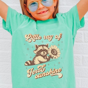 K&ouml;nnte beinhalten: Hellgr&uuml;nes T-Shirt mit dem Text "Little ray of feral sunshine" und einer Grafik eines Waschb&auml;ren, der eine Sonnenblume h&auml;lt. Das T-Shirt wird von einer Person getragen, die eine blaue Sonnenbrille und blaue Jeans tr&auml;gt.