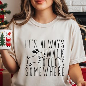 May include: A cream-colored t-shirt with the text "IT'S ALWAYS WALK O'CLOCK SOMEWHERE" and a dog graphic. The person is wearing plaid pajama pants and Christmas socks, holding a mug with a Christmas tree and Santa design.