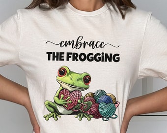 May include: White t-shirt with the text "embrace THE FROGGING" and a green frog holding a heart-shaped ball of yarn. The frog is surrounded by balls of yarn in a bowl.