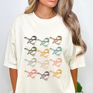 May include: A white t-shirt with a graphic of nine colorful birds in a row. The birds are all facing the same direction and have a retro style.