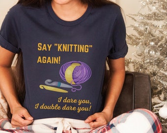 May include: A navy blue t-shirt with the text "SAY "KNITTING" AGAIN!" in yellow. The shirt also features a purple and yellow yarn ball, a crochet hook, and a knitting needle. The text "I dare you, I double dare you!" is also printed on the shirt.