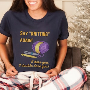 May include: A navy blue t-shirt with the text "SAY "KNITTING" AGAIN!" in yellow. The shirt also features a purple and yellow yarn ball, a crochet hook, and a knitting needle. The text "I dare you, I double dare you!" is also printed on the shirt.