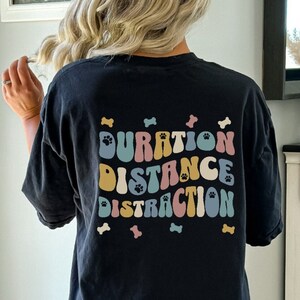 May include: Black t-shirt with a paw print on the front and the words "Duration Distance Distraction" in colorful retro font on the back. The words are surrounded by small paw prints.