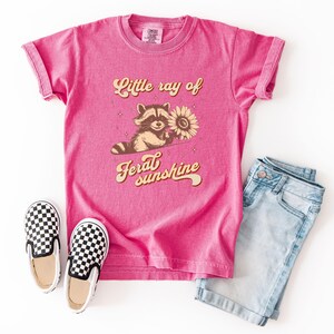K&ouml;nnte beinhalten: Ein pinkfarbenes T-Shirt mit dem Text "Little ray of feral sunshine" und einem Waschb&auml;ren, der eine Sonnenblume h&auml;lt. Dazu eine hellblaue Jeansshorts und schwarz-wei&szlig; karierte Slip-on-Schuhe.