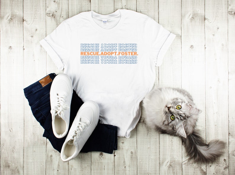 May include: White t-shirt with the text "RESCUE.ADOPT.FOSTER." printed in blue and orange on a white background. The shirt is laid out on a wooden surface with a pair of white sneakers and blue jeans. A gray cat is lying on the surface.