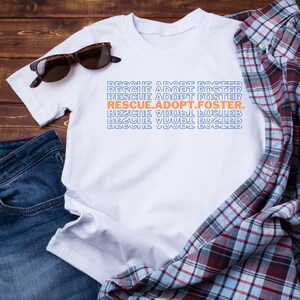 May include: White t-shirt with the text "RESCUE.ADOPT.FOSTER." repeated in blue and orange. The shirt is laid flat on a wooden surface with a pair of sunglasses and a blue denim shirt.