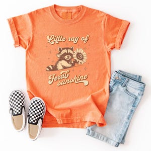 K&ouml;nnte beinhalten: Orangefarbenes T-Shirt mit hochgekrempelten &Auml;rmeln mit einem Waschb&auml;ren, der eine Sonnenblume h&auml;lt, mit dem Text "Little ray of feral sunshine". Das Shirt wird mit hellblauen Jeansshorts und schwarz-wei&szlig; karierten Slip-On-Schuhen kombiniert.