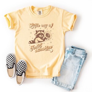 K&ouml;nnte beinhalten: Ein gelbes T-Shirt mit einem Waschb&auml;r-Motiv, das eine Sonnenblume h&auml;lt, und dem Text "Little ray of feral sunshine". Das Shirt wird mit hellblauen Jeansshorts und schwarz-wei&szlig; karierten Slip-On-Schuhen kombiniert.