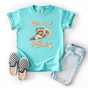 K&ouml;nnte beinhalten: T&uuml;rkisfarbenes T-Shirt mit dem Text "Little ray of feral sunshine" und einer Grafik eines Waschb&auml;ren, der eine Sonnenblume h&auml;lt. Das Bild zeigt Jeansshorts und schwarz-wei&szlig; karierte Slip-on-Schuhe.