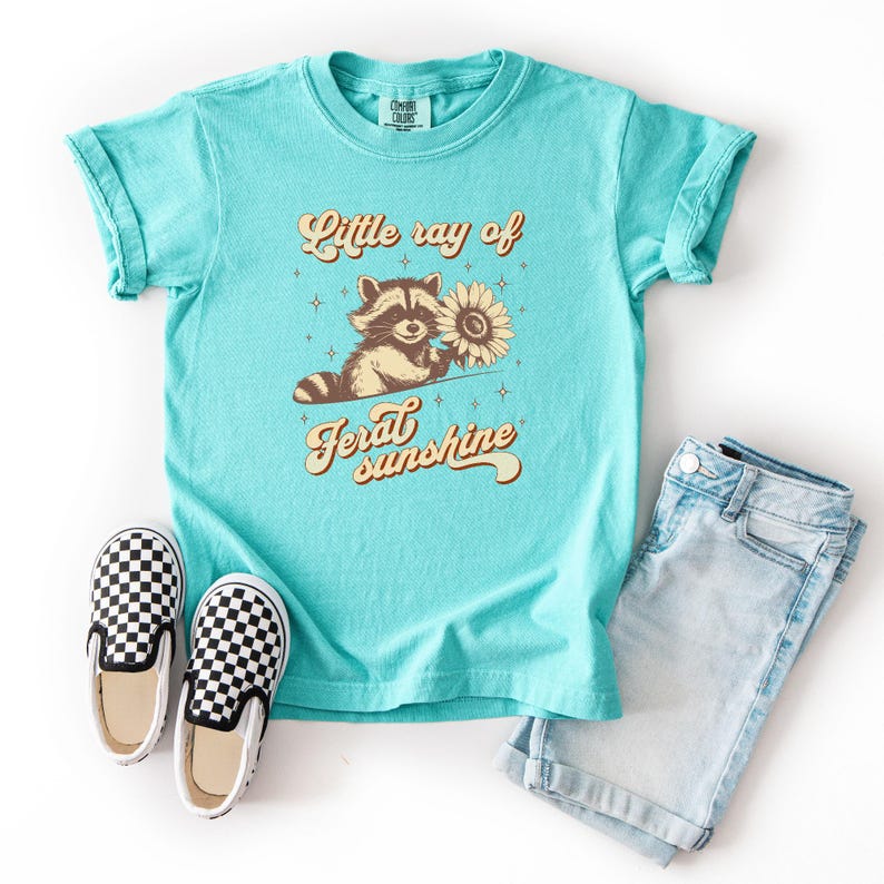 K&ouml;nnte beinhalten: T&uuml;rkisfarbenes T-Shirt mit dem Text "Little ray of feral sunshine" und einer Grafik eines Waschb&auml;ren, der eine Sonnenblume h&auml;lt. Das Bild zeigt Jeansshorts und schwarz-wei&szlig; karierte Slip-on-Schuhe.