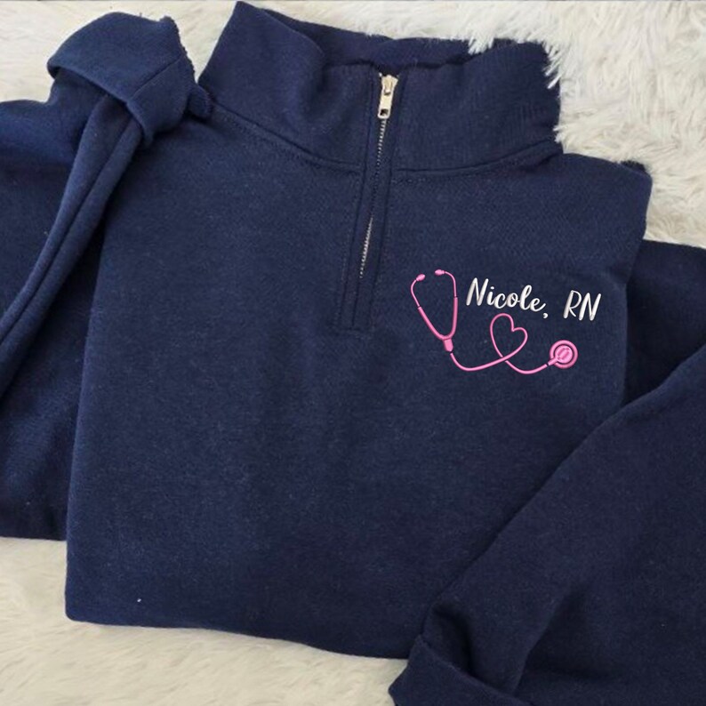 May include: Navy blue quarter-zip sweatshirt with a pink stethoscope and heart design and the text "Nicole, RN".