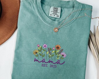 May include: A teal blue t-shirt with a floral design and the text "mama est. 2023" in purple.