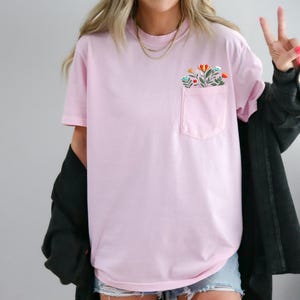 May include: A pink t-shirt with a floral embroidered pocket. The shirt has a relaxed fit and short sleeves.