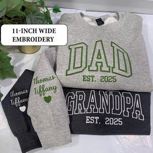 May include: A light gray sweatshirt with green embroidered text reading DAD EST. 2025 and a dark gray sweatshirt with white embroidered text reading GRANDPA EST. 2025. Sleeves feature embroidered names Thomas and Tiffany with a small heart.