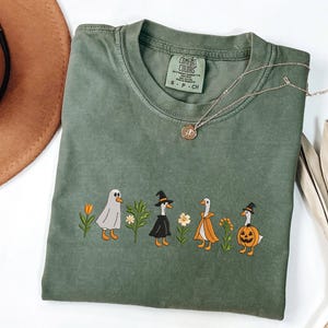 May include: An olive green t-shirt featuring an embroidered graphic of four geese in Halloween costumes, including a ghost, a witch, a cape, and a pumpkin, interspersed with small colorful flowers. The shirt has a Comfort Colors brand label.