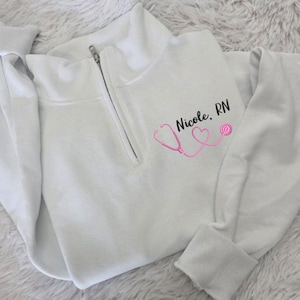 May include: White 1/4 zip sweatshirt with a pink stethoscope and two hearts design. The text "Nicole, RN" is embroidered on the sweatshirt.