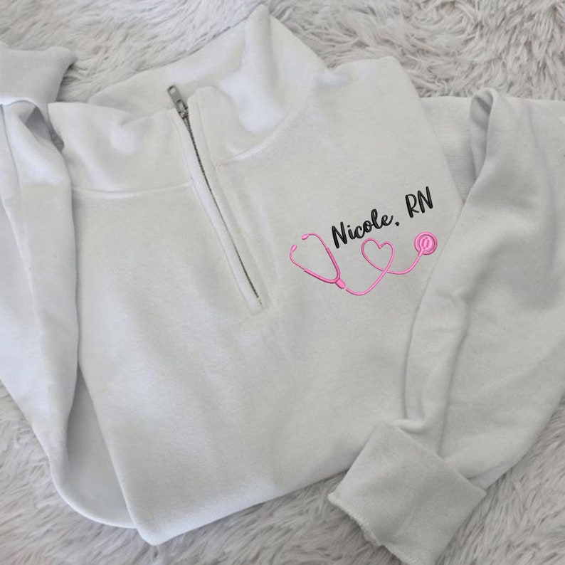 May include: White 1/4 zip sweatshirt with a pink stethoscope and two hearts design. The text "Nicole, RN" is embroidered on the sweatshirt.
