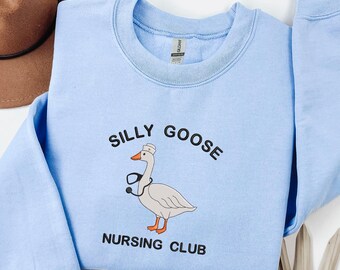 May include: A light blue sweatshirt with the words "SILLY GOOSE NURSING CLUB" printed in black. The graphic features a cartoon goose wearing a nurse's cap and stethoscope. A brown hat is visible in the background.