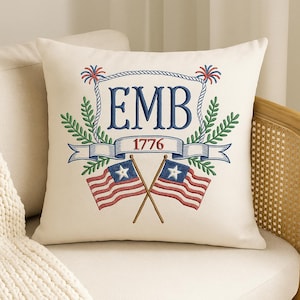 May include: Cream-colored throw pillow with an embroidered patriotic design. Features the initials "EMB" in blue, the year "1776", crossed American flags, and decorative elements in red, blue, and green. The pillow is on a white chair.