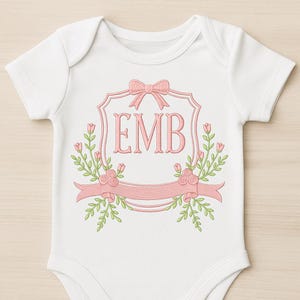 May include: White baby onesie with a pink floral design. The design includes a pink bow, the letters "EMB", and a pink banner with floral accents. The onesie is made of soft material.