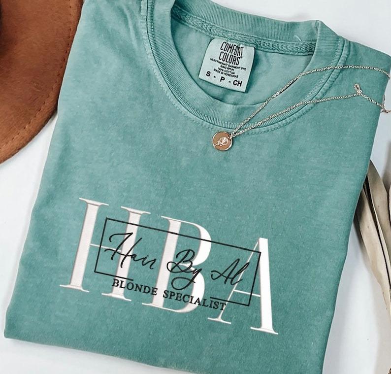 May include: A teal blue t-shirt with the text "Hair By Al" in a white box and the words "Blonde Specialist" underneath. The shirt is made of a soft, comfortable material.