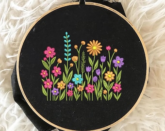 May include: Embroidered hoop art featuring a colourful floral design with pink, purple, orange, yellow, blue, and green flowers on a black background.