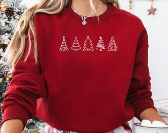 May include: A red sweatshirt featuring a white line drawing of five Christmas trees. The trees vary in design, with stars atop some. The sweatshirt is part of a holiday-themed clothing collection.