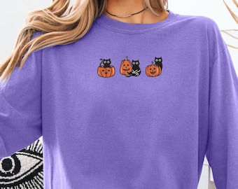 May include: A purple sweatshirt featuring a Halloween-themed design. The design includes three orange pumpkins with black cat faces. The sweatshirt is a solid purple color and has a crew neck.