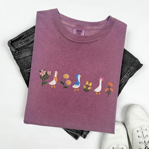 May include: A mauve colored t-shirt with an embroidered design of five ducks in a row. The ducks are white with different colored details, and there are flowers and leaves embroidered around them. The t-shirt has the text "COMFORT COLORS" on the tag.