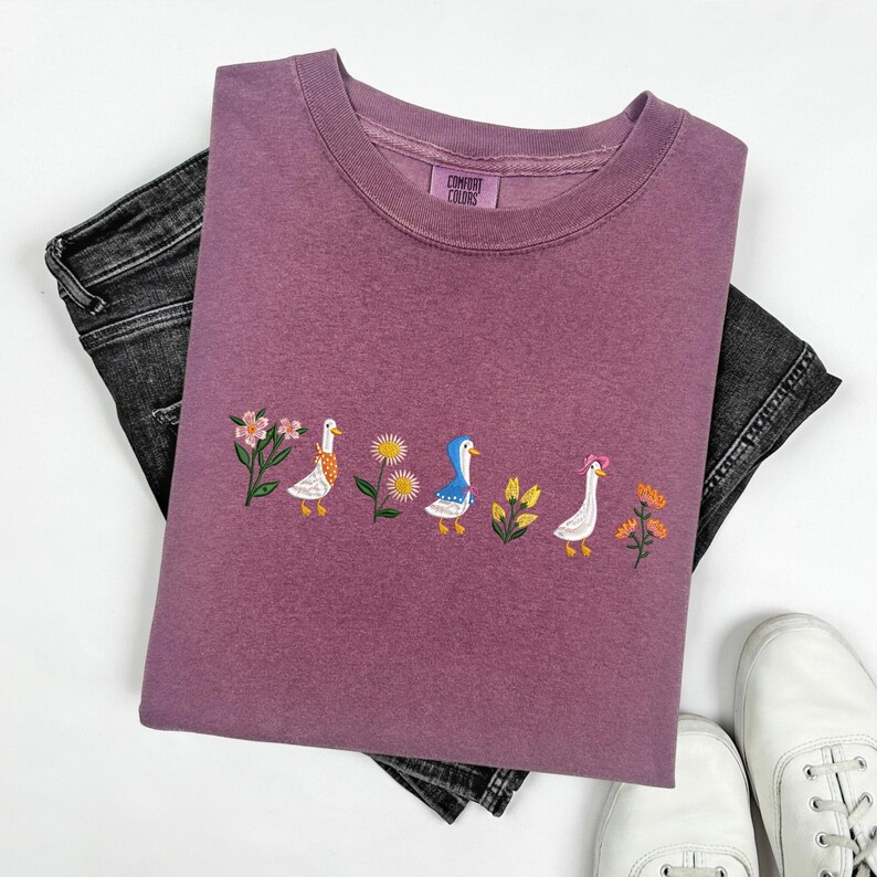 May include: A mauve colored t-shirt with an embroidered design of five ducks in a row. The ducks are white with different colored details, and there are flowers and leaves embroidered around them. The t-shirt has the text "COMFORT COLORS" on the tag.