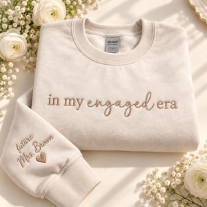 May include: A sand-colored crewneck sweatshirt featuring the embroidered text "in my engaged era" across the chest in a brown script font. The sleeve is embroidered with the text "future Mrs. Brown" and a small brown heart icon.