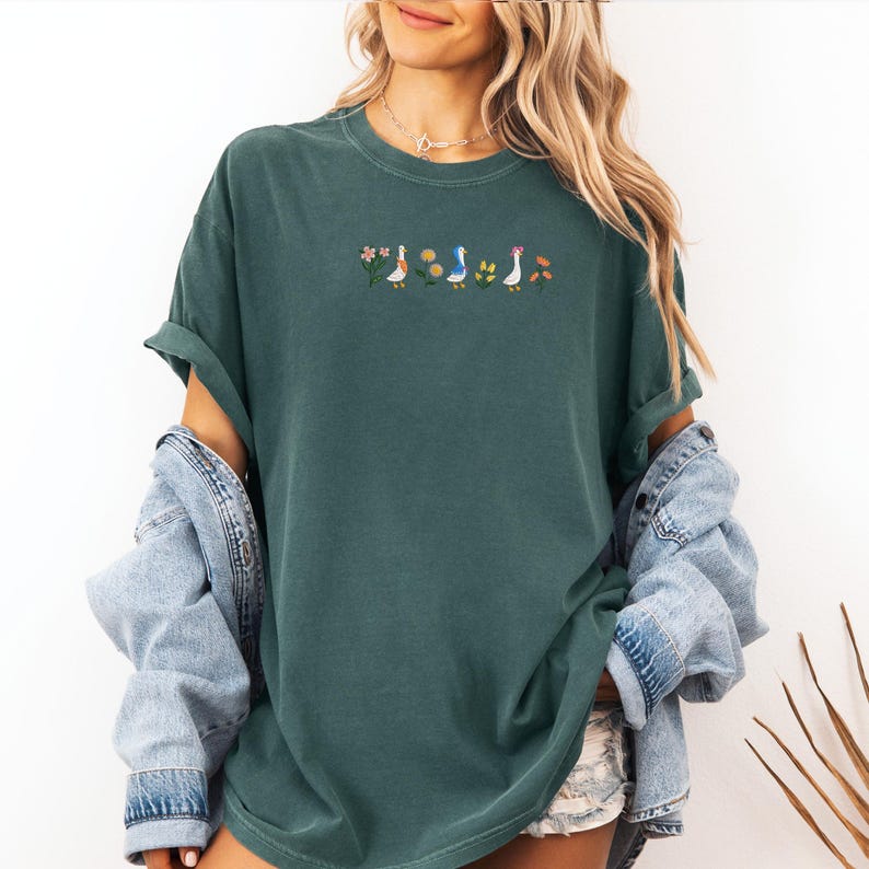 May include: A dark green t-shirt with a line of embroidered ducks and flowers. The ducks are wearing different colored hats.